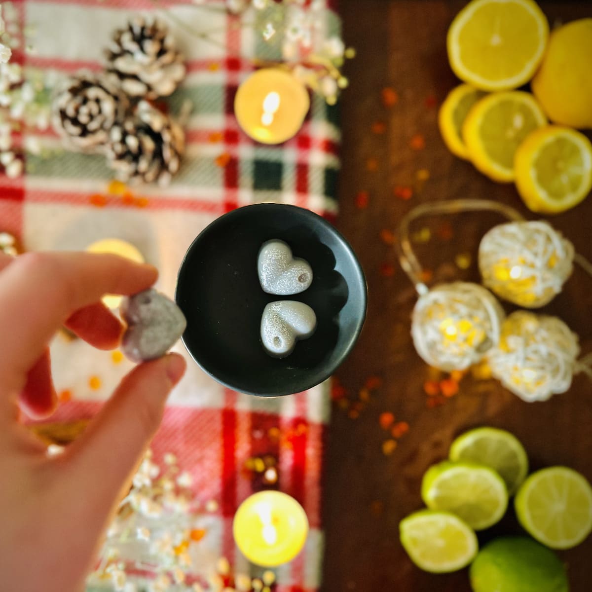 Hand holding a grey Starry Night heart shaped wax melt and placing into a black tealight burner dish wish a Christmas background surrounded by lemons, limes and tealights