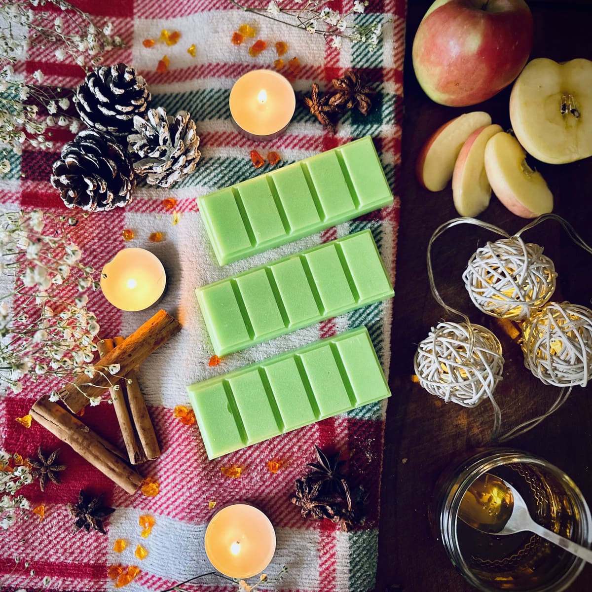 Green  coloured Apple and Cinnamon scented snapbar wax melts on a festive checkered cloth with tealights, apples, cinnamon and christmas lights.