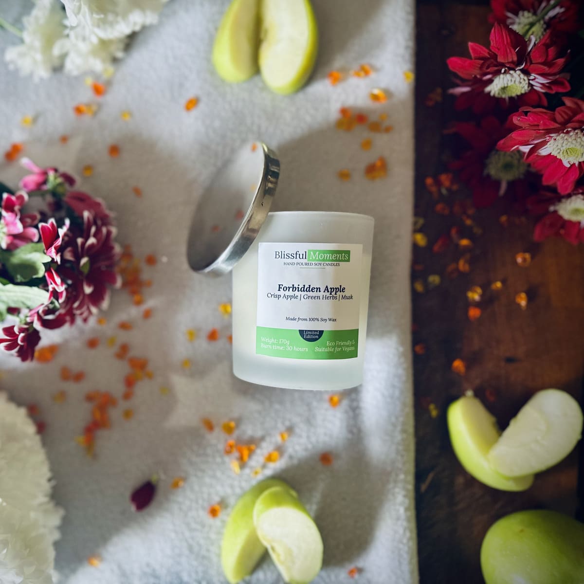 Forbidden Apple scented luxury frosted glass soy candle surrounded by fresh flowers, amber and green apples sitting on a cosy blanket