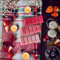 Red coloured Frosty Berries snapbar wax melts on a checked plaid cloth with candles, winter berries, and fruits.