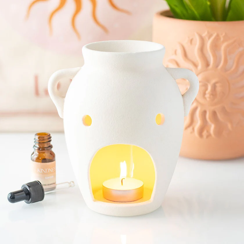 White greek vase inspired ceramic wax melt burner with a lit candle inside, surrounded by a bottle of essential oil and a plant.