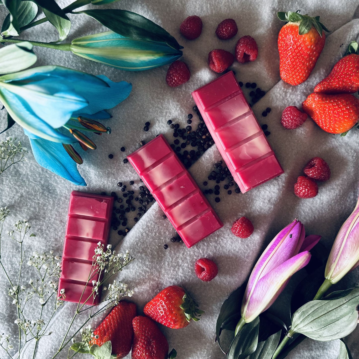 Red & white soy wax melts surrounded by fresh strawberries and lilies sitting on black peppercorns and a wooly blanket