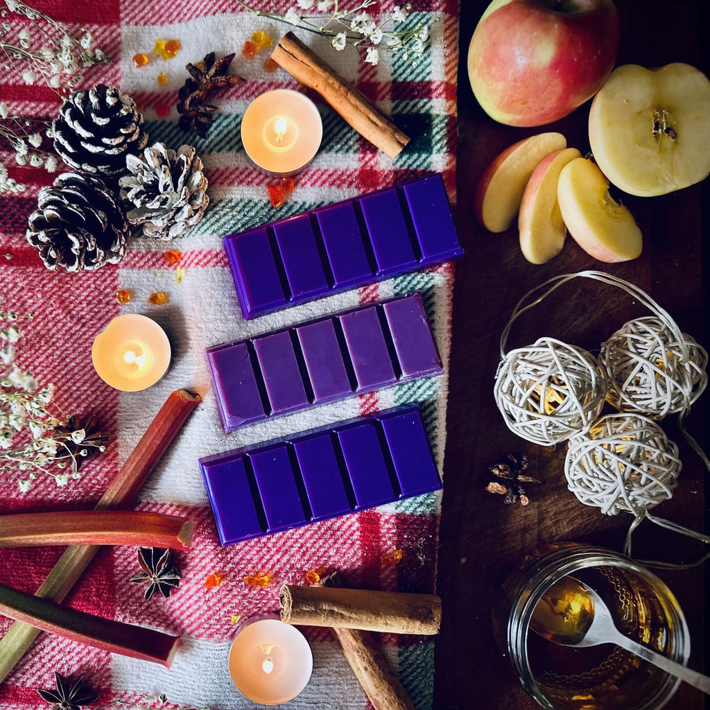 Purple coloured Rhubarb and Cinnamon scented snapbar wax melts on a festive red checkered cloth with tea lights, apples, cinnamon and decorative elements.