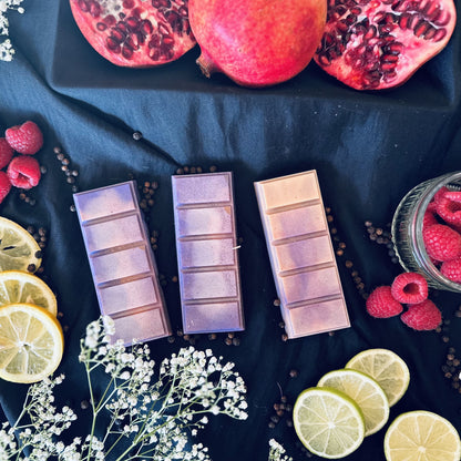 Orange and purple soy wax melts sitting on a bed of peppercorns surrounded by pomegranates,  fresh raspberries an citrus fruits