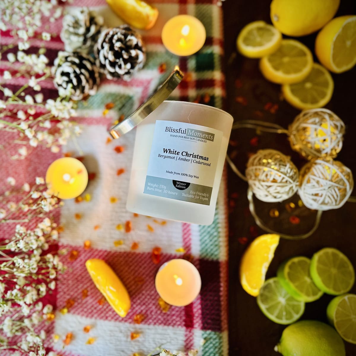 White Christmas scented soy candle on a checkered tablecloth with tea lights, lemons, and limes in a Christmas decorative setting
