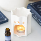 White ceramic wax melt burner shaped as a star with a lit candle inside, surrounded by books