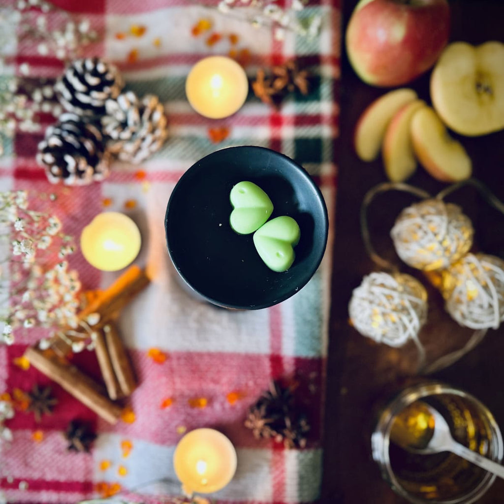 Green coloured Apple and Cinnamon scented heart shaped wax melts in a black wax melt burner on a festive plaid fabric background with tea lights and apples, cinnamon and caramel.