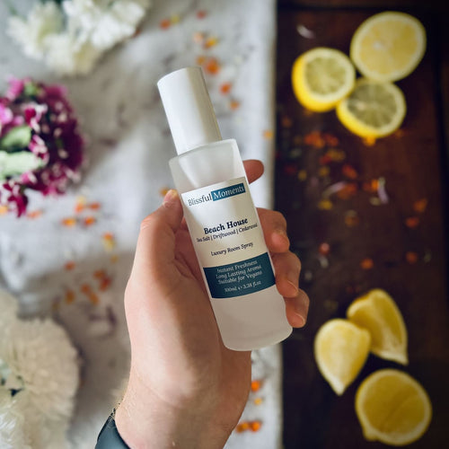 Hand holding Beach House scented luxury frosted glass room spray surrounded by fresh flowers, amber and lemon