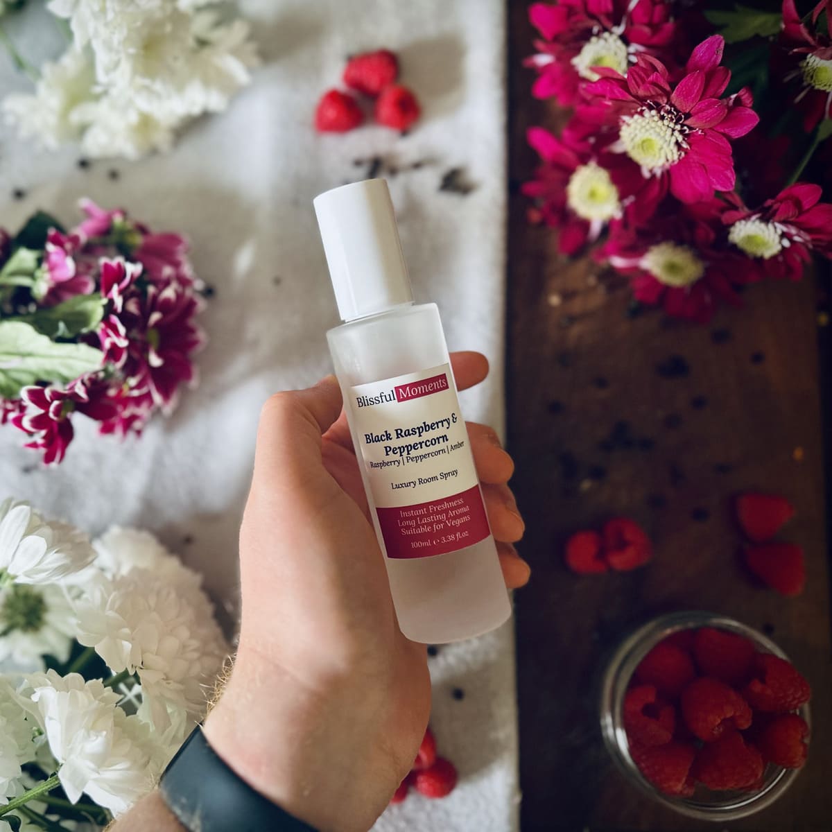 Hand holding Black Raspberry & Peppercorn scented luxury frosted glass room spray surrounded by fresh flowers, peppercorns and raspberries sitting on a cosy blanket