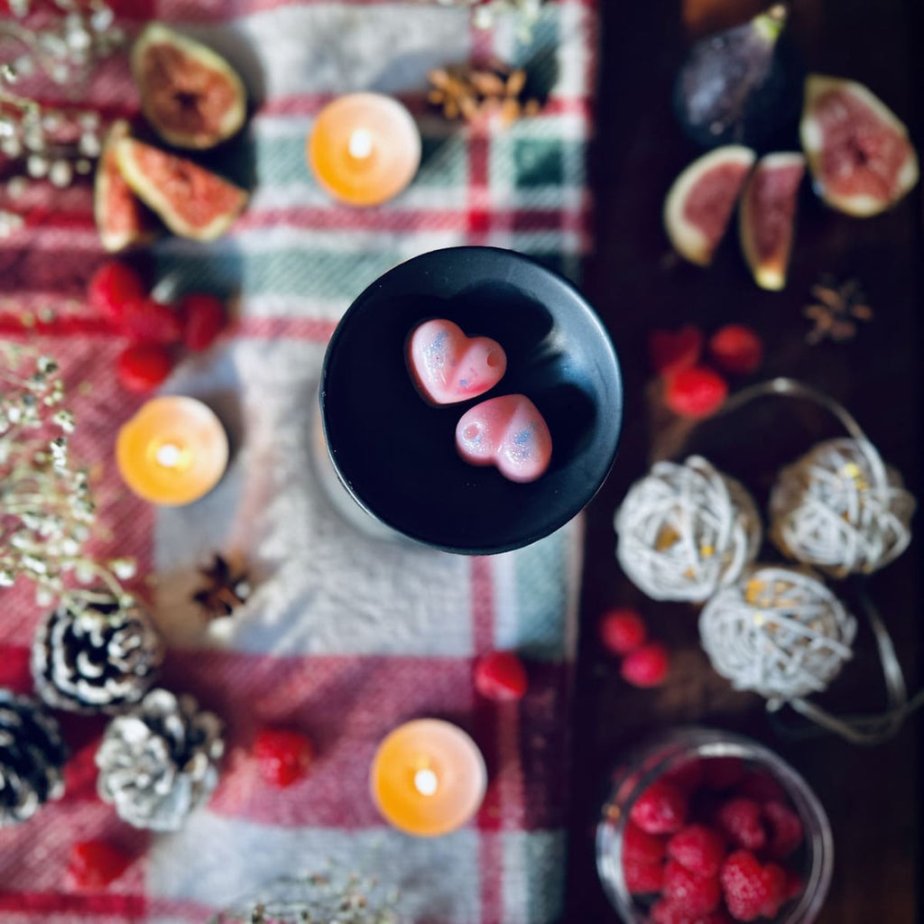 Pink coloured Cherry and Fig scented heart shaped wax melts in a black wax melt burner on a festive red checked tablecloth surrounded by tea lights, figs, and pinecones