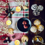 Red coloured Christmas Cookie scented heart shaped wax melts in a black wax melt burner on a festive tablecloth surrounded by chestnuts, vanilla, biscuits and caramel.