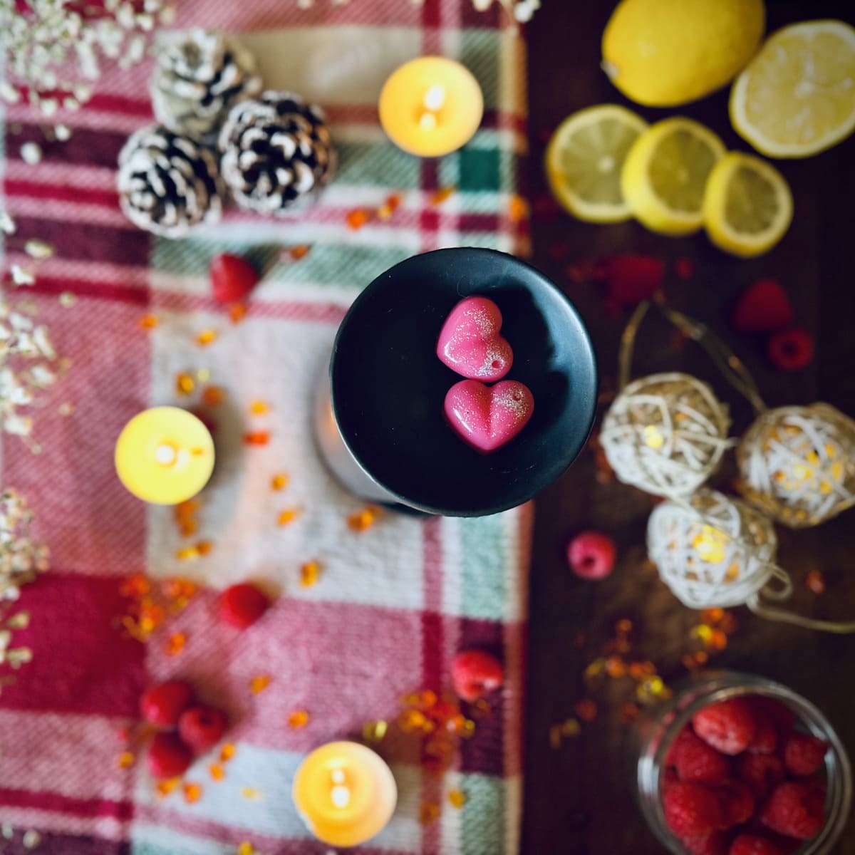 Red Christmas scented heart shaped wax melts in a black wax melt burner dish in Decorative setting with candles, fruits, and a plaid cloth