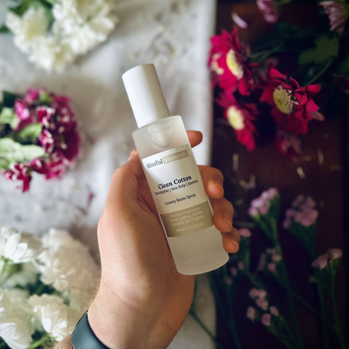 Hand holding a luxury Clean Cotton scented room spray surrounded by fresh flowers.