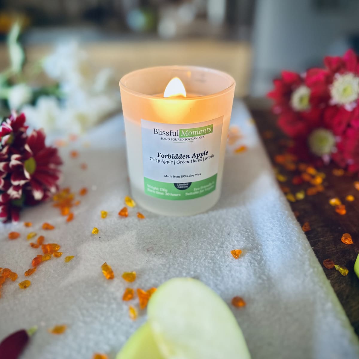 Forbidden Apple scented luxury frosted glass soy candle surrounded by fresh flowers, amber and green apples sitting on a cosy blanket