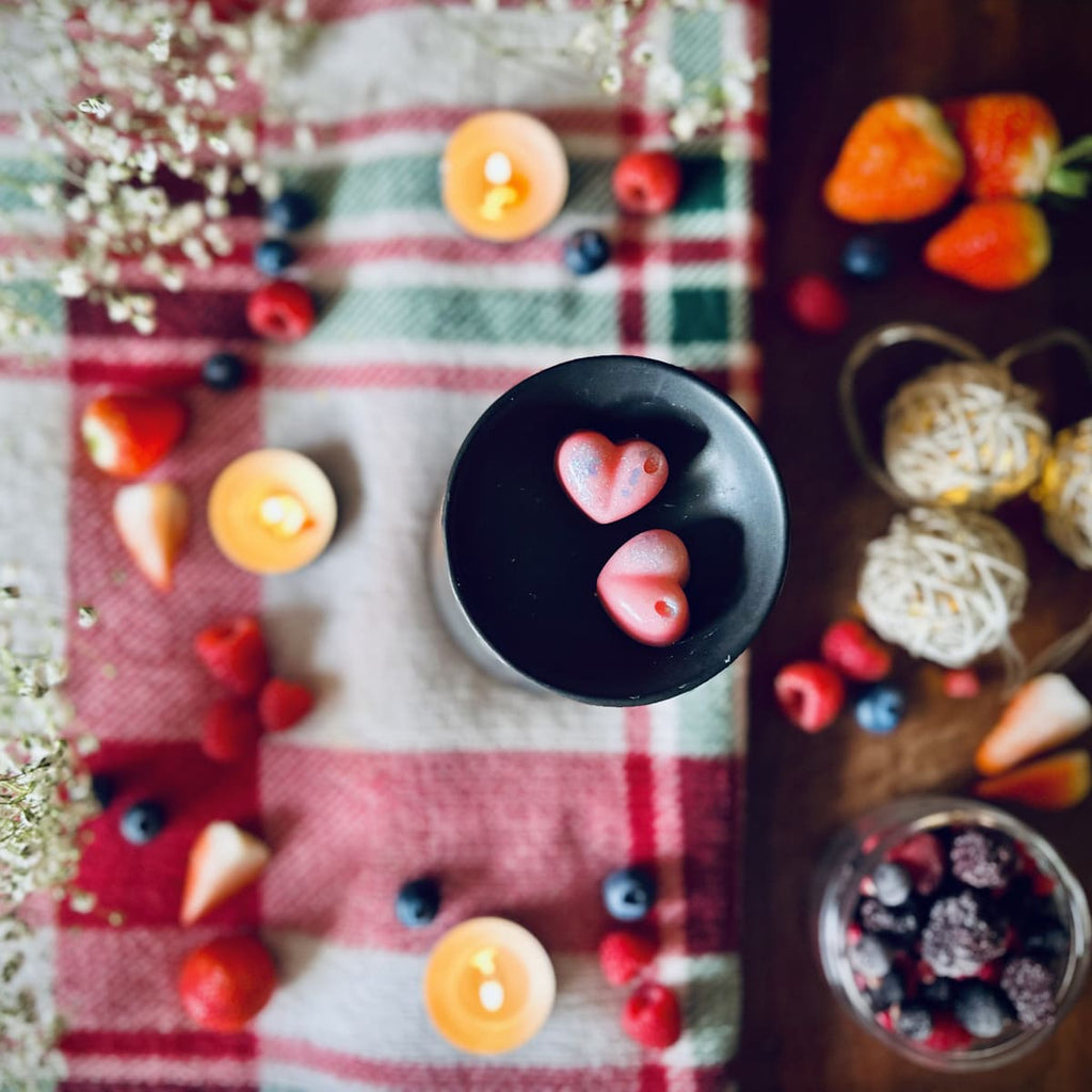 Pink coloured Frosty Berried scented heart shaped wax melts in a black wax melt burner dish in Decorative festive setting with candles, winter berries, and a plaid cloth.