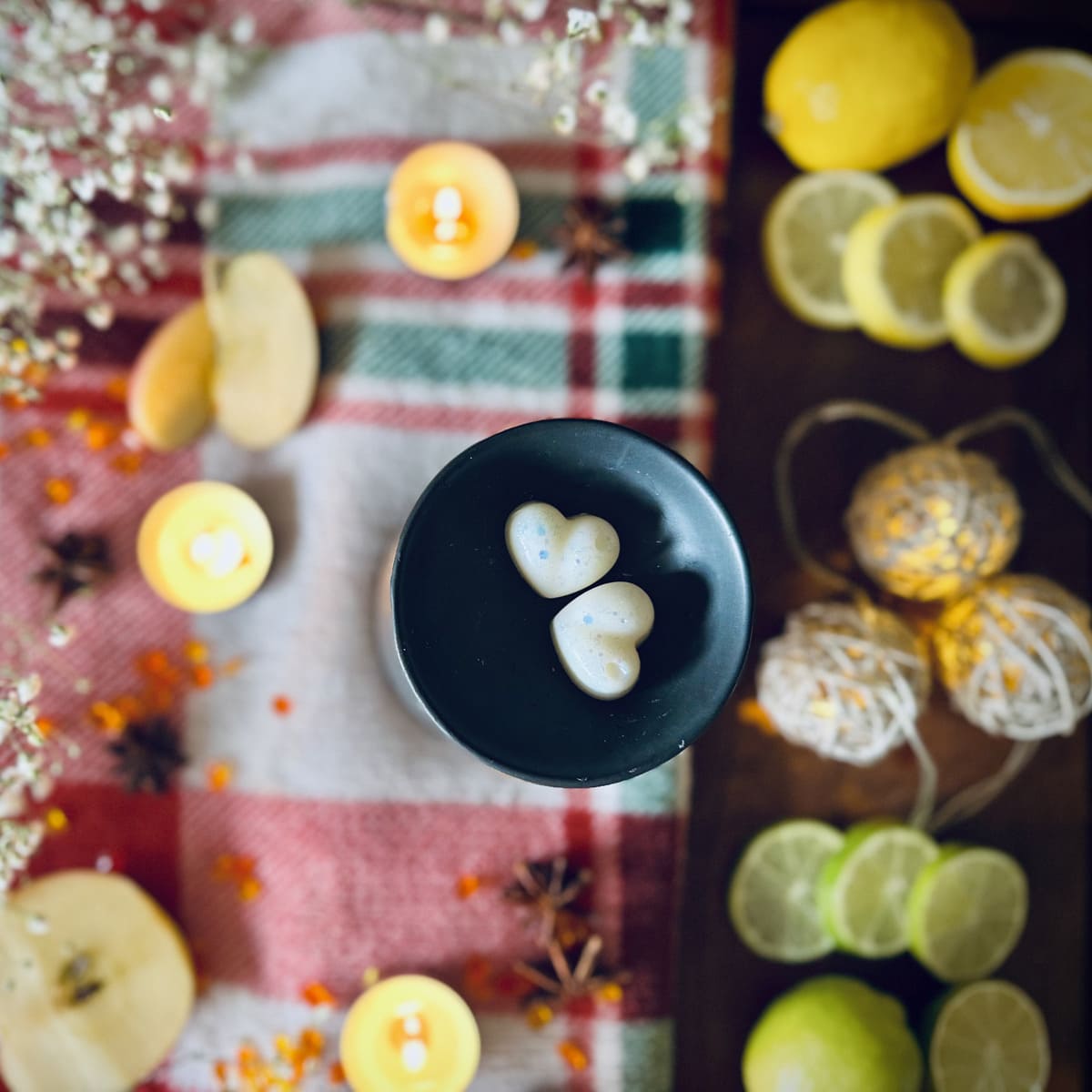 Two white coloured Frosty Lake scented heart shaped wax melts in a black wax melt burner dish on a festive tablecloth surrounded by tea lights with candles and citrus fruits and Christmas lights