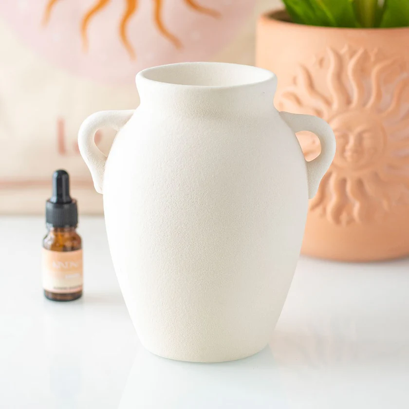 White greek vase inspired ceramic wax melt burner with a lit candle inside, surrounded by a bottle of essential oil and a plant.
