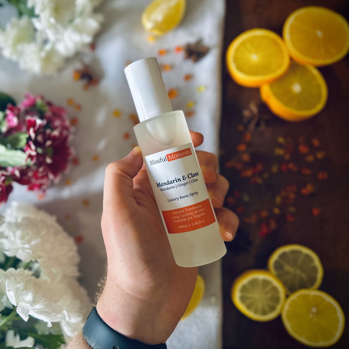 Hand holding a luxury Mandarin & Clove scented frosted glass room spray surrounded by fresh flowers and citrus rooms sitting on a cosy blanket