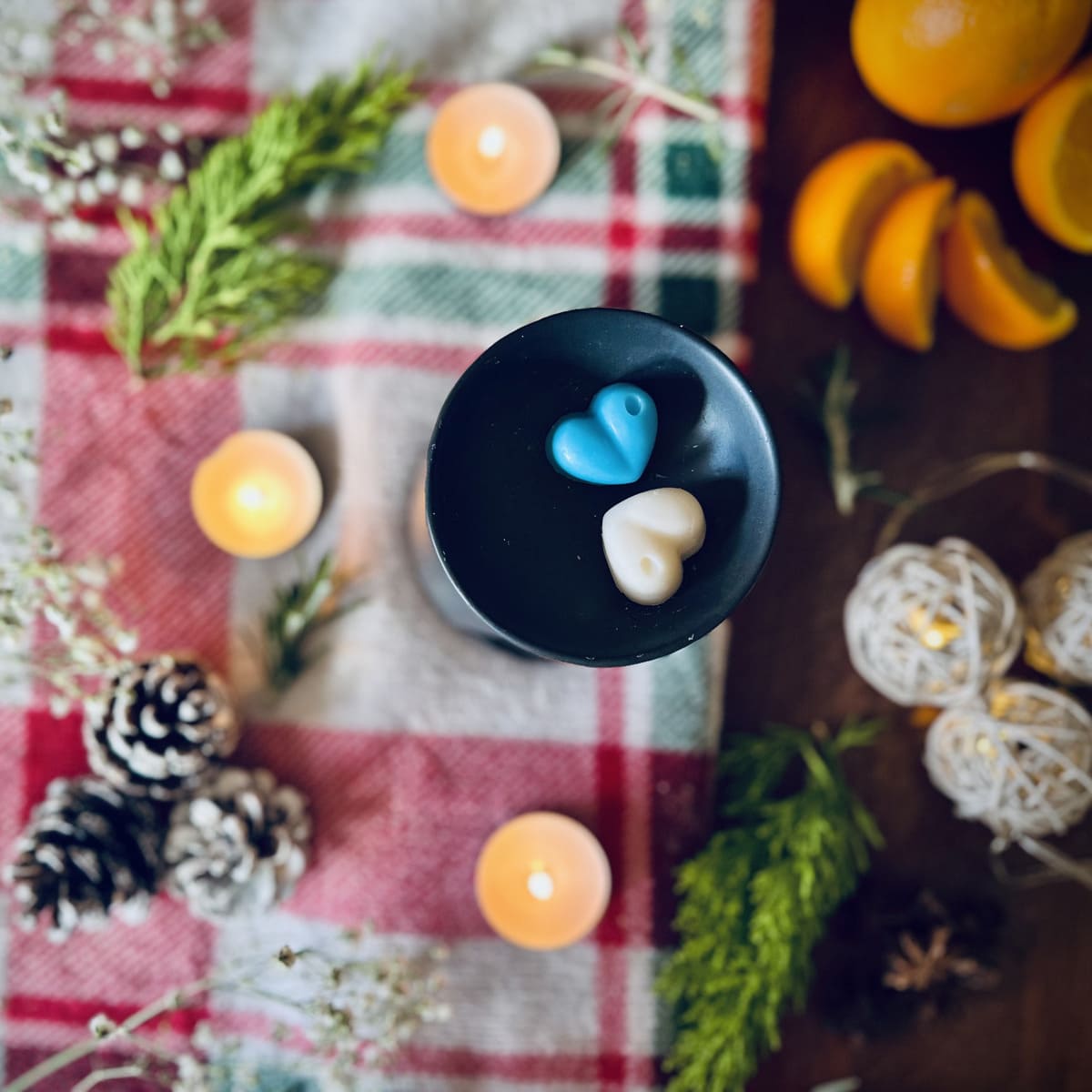 Blue and white coloured Northern lighted scented heart shaped wax melts in a black wax melt burner on a festive tablecloth with tea lights and oranges and rosemary.