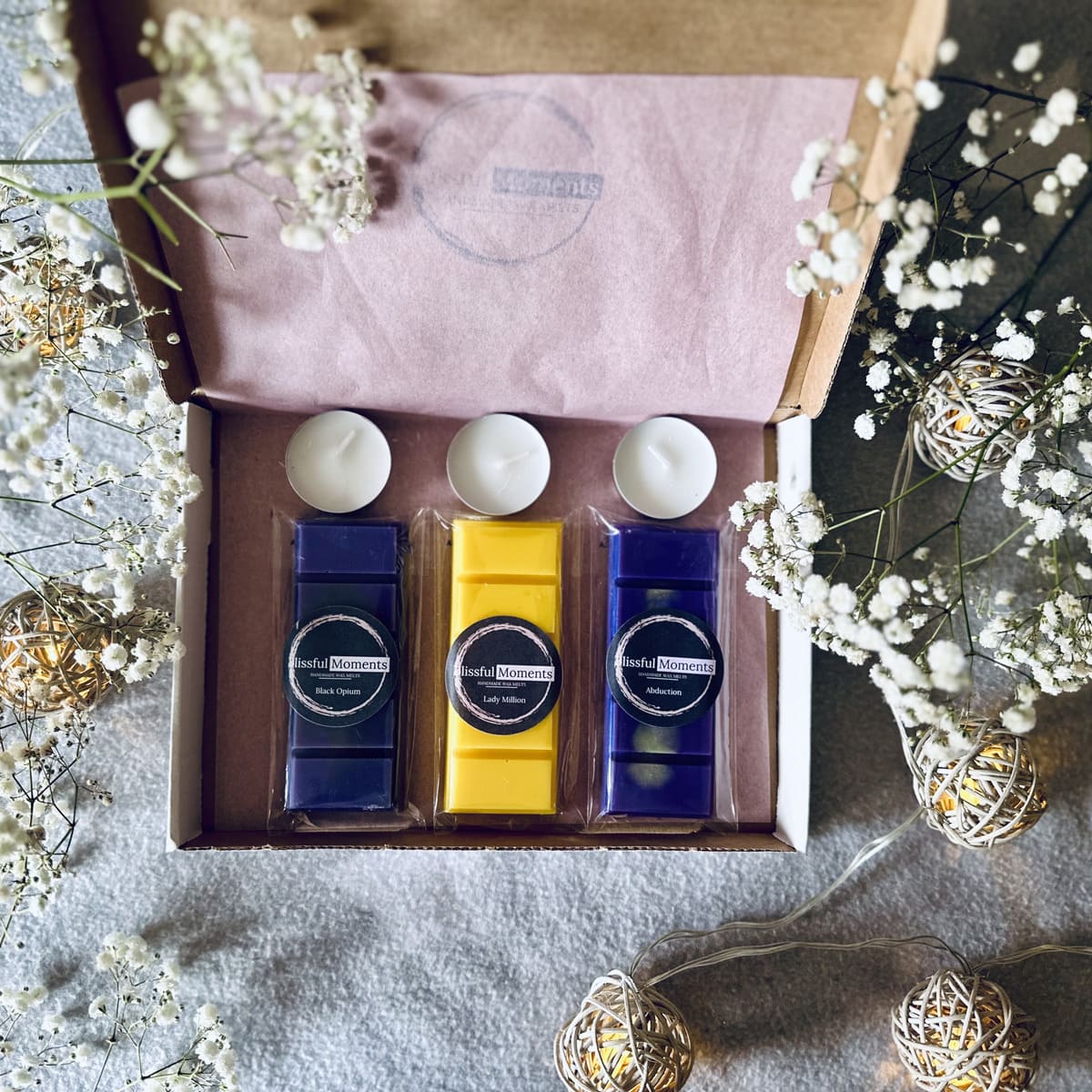 Luxurious purple and gold coloured soy welts in a gift box sitting next to tea lights and surrounded by white flowers and fairy lights