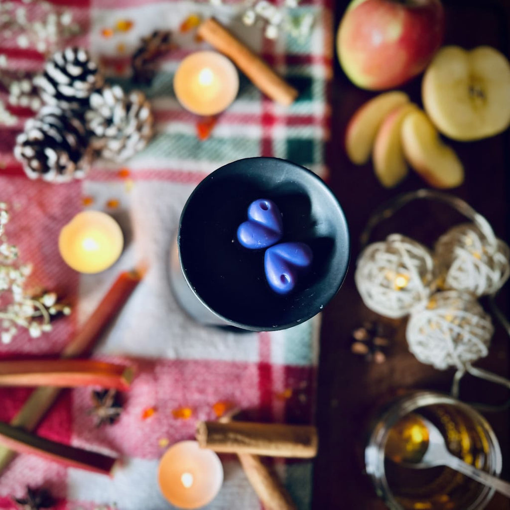 Purplee coloured Rhubarb and cinnamon scented heart shaped wax melts in a black wax melt burner on a festive table with tea lights and apples and caramel.