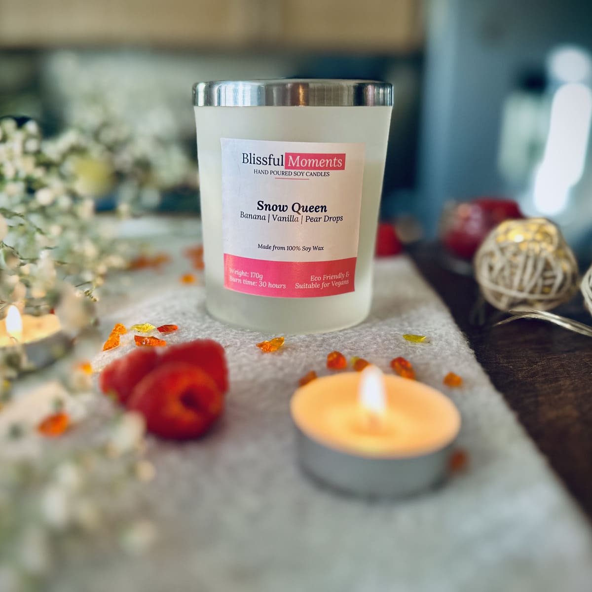 Snow Queen candle with a silver lid on a grey blanket surrounded flowers, tea lights and raspberries