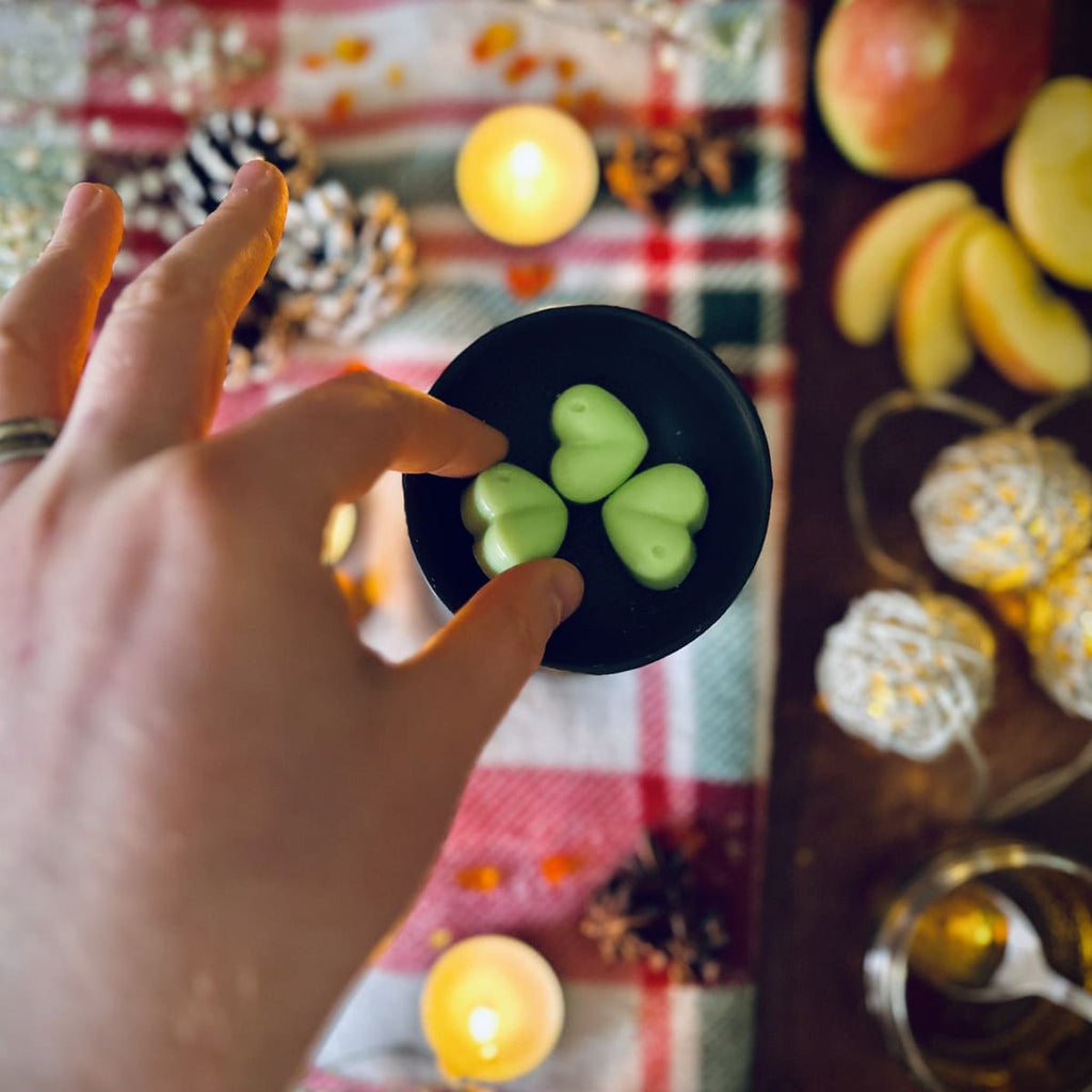Hand holding a green coloured Apple and Cinnamon scented heart shaped wax melt against a festive checked tablecloth background with tea lights and apples, cinnamon and caramel.