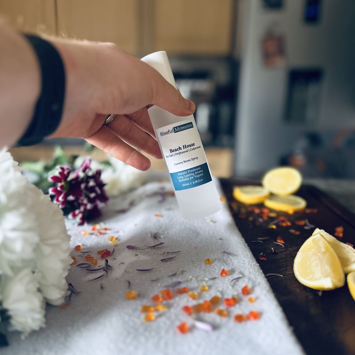 Hand holding Beach House scented luxury frosted glass room spray surrounded by fresh flowers, amber and lemon