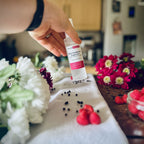 Hand holding Black Raspberry & Peppercorn scented luxury frosted glass room spray surrounded by fresh flowers, peppercorns and raspberries 