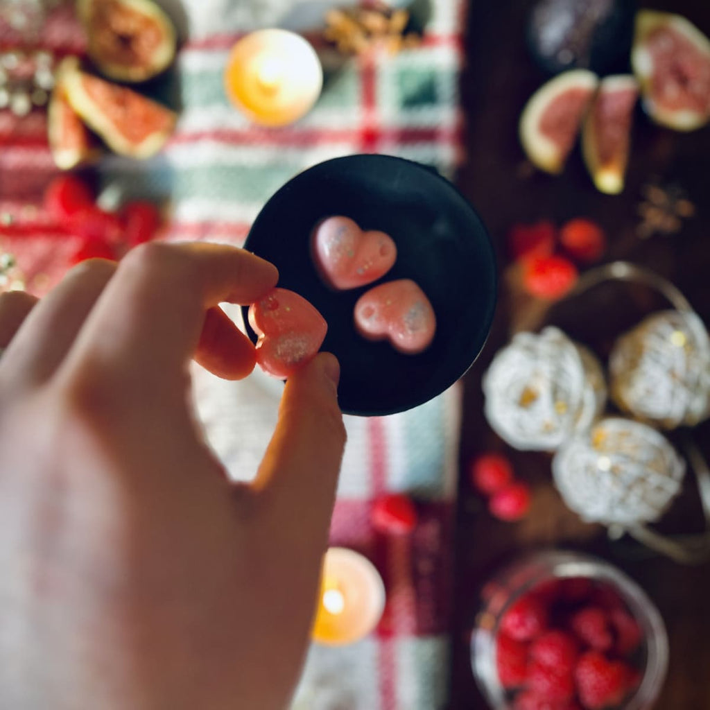 Hand holding a pink coloured cherry and fig scented heart shape wax melt on.a Christmas tablecloth with festive items