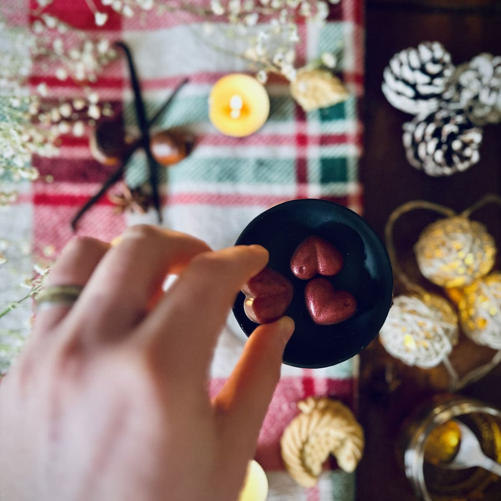 A Hand holding a red Christmas Cookie scented heart shaped wax melts  against a festive background with chestntuts, vanilla and caramel