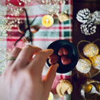 A Hand holding a red Christmas Cookie scented heart shaped wax melts  against a festive background with chestntuts, vanilla and caramel