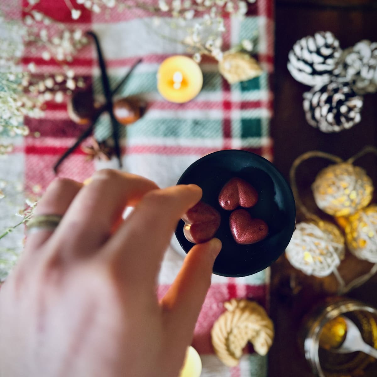 A Hand holding a red Christmas Cookie scented heart shaped wax melts  against a festive background with chestntuts, vanilla and caramel