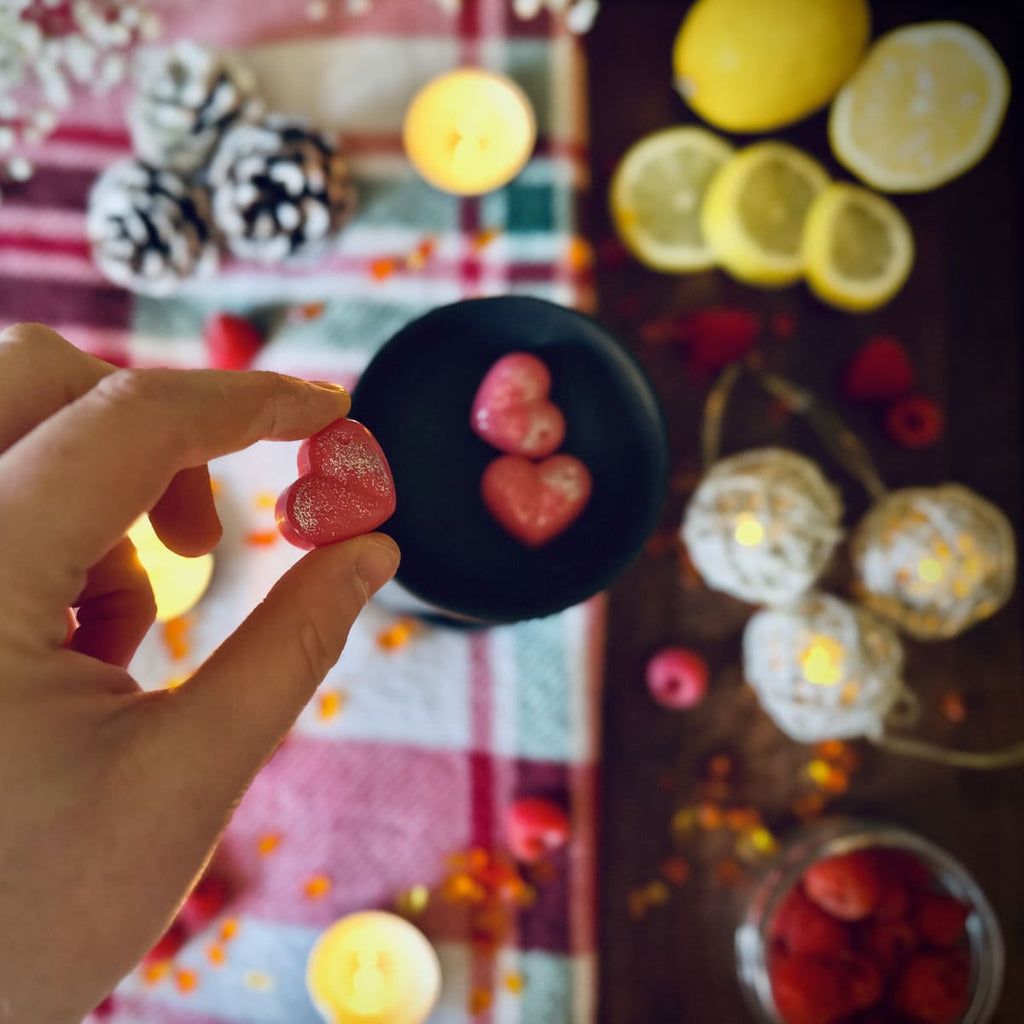 Hand holding a red Christmas Wish scented heart shaped wax melt with a blurred festive background featuring lemons, berries, and lights.
