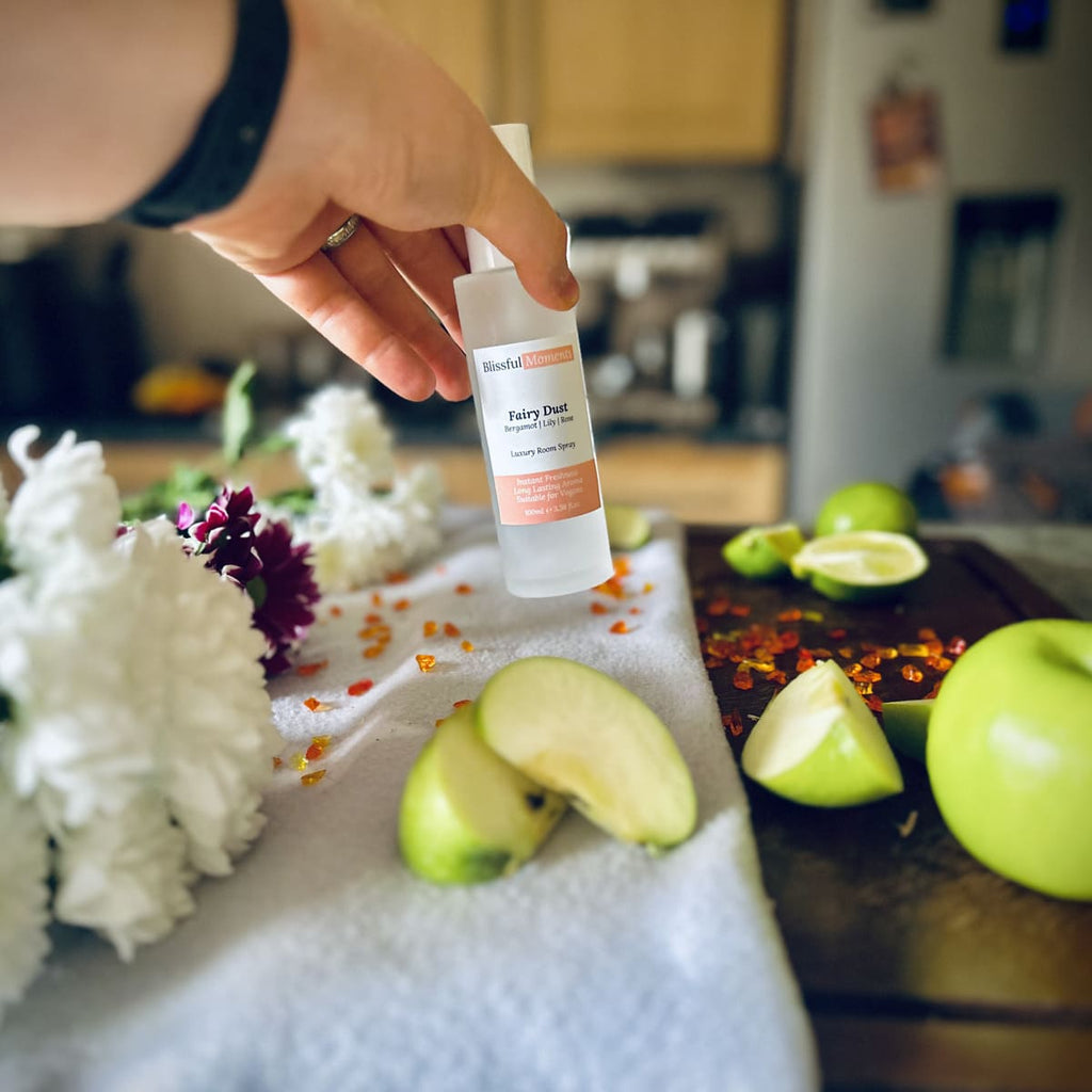 Hand holding Fairy Dust scented luxury frosted glass room spray surrounded by fresh flowers, amber and green apples sitting on a cosy blanket