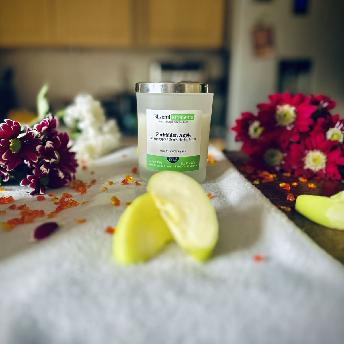 Forbidden Apple scented luxury frosted glass soy candle surrounded by fresh flowers, amber and green apples sitting on a cosy blanket