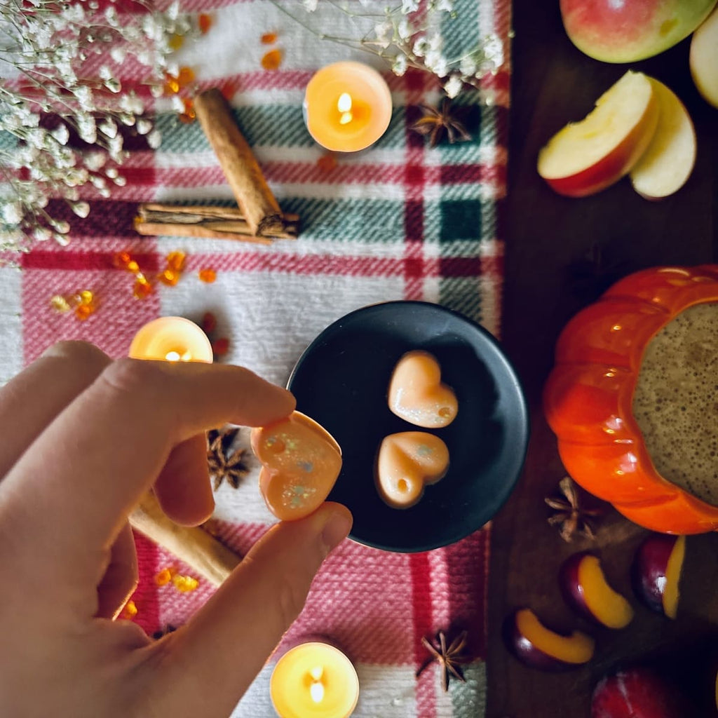 Hand holding a Orange Frosted Pumpkin scented heart shaped wax melts on a red plaid tablecloth with candles and apples, plumbs and cinnamon