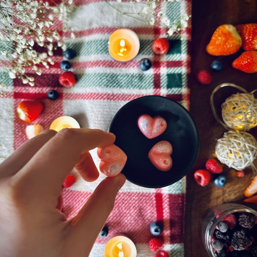 A Hand holding a black pink coloured Frosty Berries scented heart shaped wax melt in a festive decorative setting on tablecloth with winter berries and tealights.