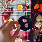 A Hand holding a black pink coloured Frosty Berries scented heart shaped wax melt in a festive decorative setting on tablecloth with winter berries and tealights.
