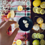 A Hand holding a white coloured Frosty Lake scented heart shaped wax melt over a festive background with lemons and Christmas lights.