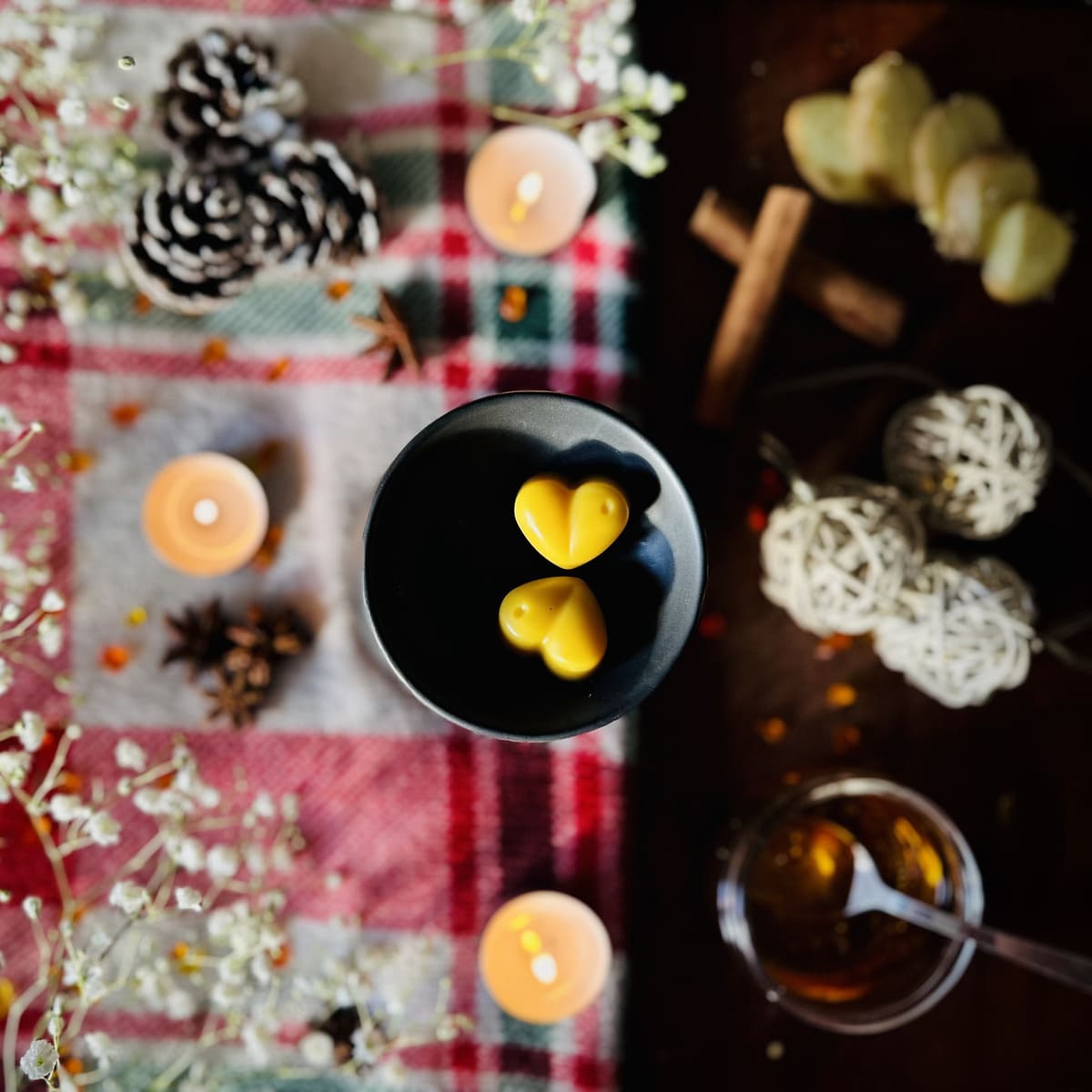 Golden Gingerbread Scented heart-shaped wax melts sitting in a black tealight burner disk surrounded by candles and decorative Christmas decoration on a plaid tablecloth.