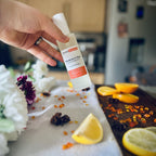 Hand holding a luxury Mandarin & Clove scented frosted glass room spray surrounded by fresh flowers and citrus rooms sitting on a cosy blanket