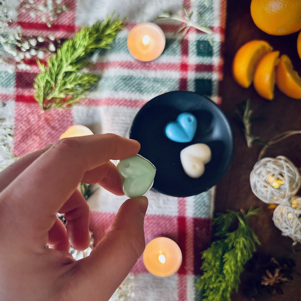 Hand holding a green coloured Northern lights heart shaped wax melt with a bue and white wax melt in a black wax melt burner on a festive background with candles and oranges.