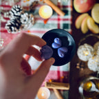 Hand holding purple coloured rhubarb and cinnamon scented heart shaped wax melt against a festive background with tea lights, apples and cinnamon sticks.