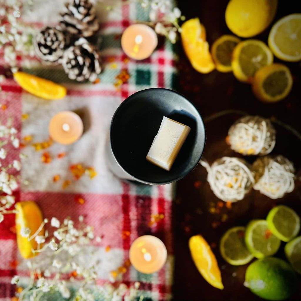 White Christmas snapbar wax melt segment sitting in a black tealight burner dish surrounded in a decorative Christmas set up with lemons, limes, and candles on a festive checkered tablecloth