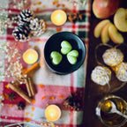 Three Green coloured Apple and Cinnamon scented heart shaped wax melts in a black wax melt burner dish on a festive table with tea lights, apples, and cinnamon sticks.
