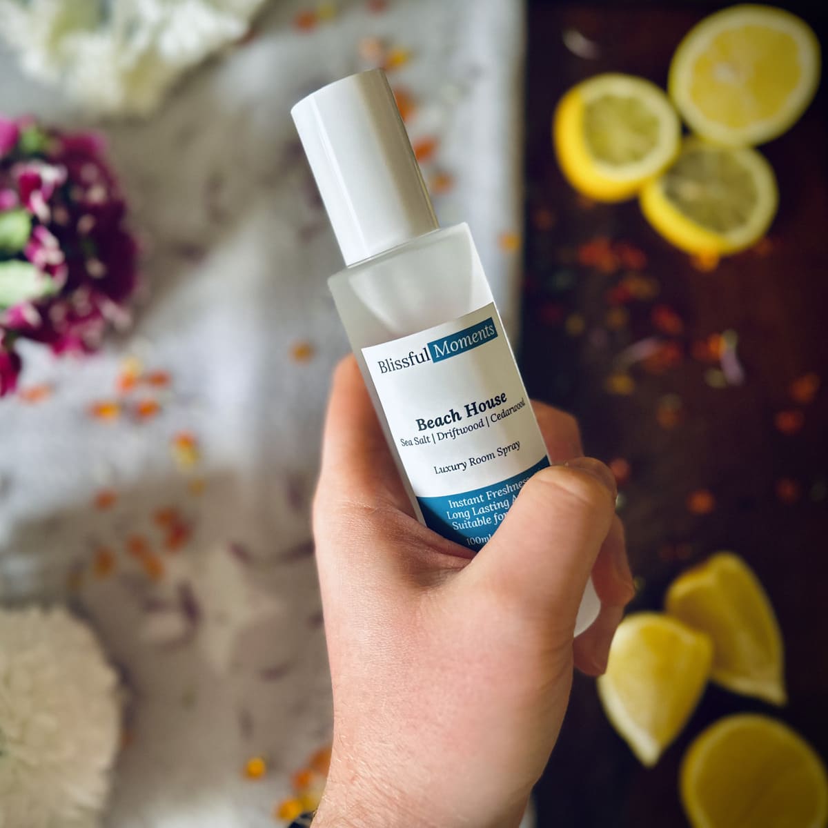 Hand holding Beach House scented luxury frosted glass room spray surrounded by fresh flowers, amber and lemon