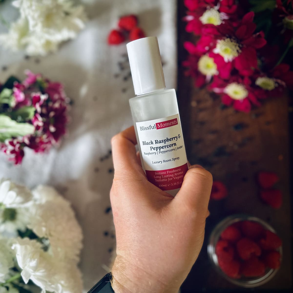 Hand holding Black Raspberry & Peppercorn scented luxury frosted glass room spray surrounded by fresh flowers, peppercorns and raspberries