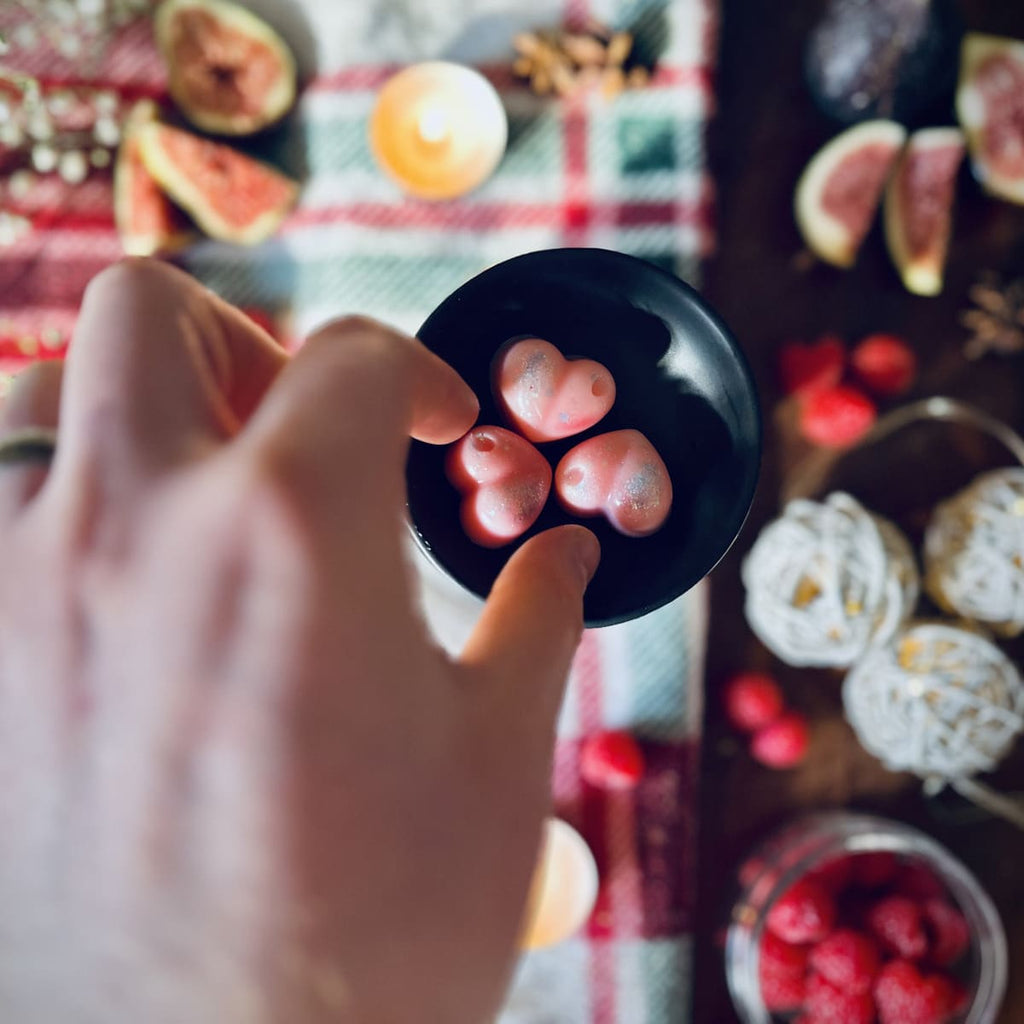 Three pink Cherry and Fig scented heart shaped wax melts in a black wax melt burner on a red festive checked tablecloth surrounded by figs, raspberries and christmas lights