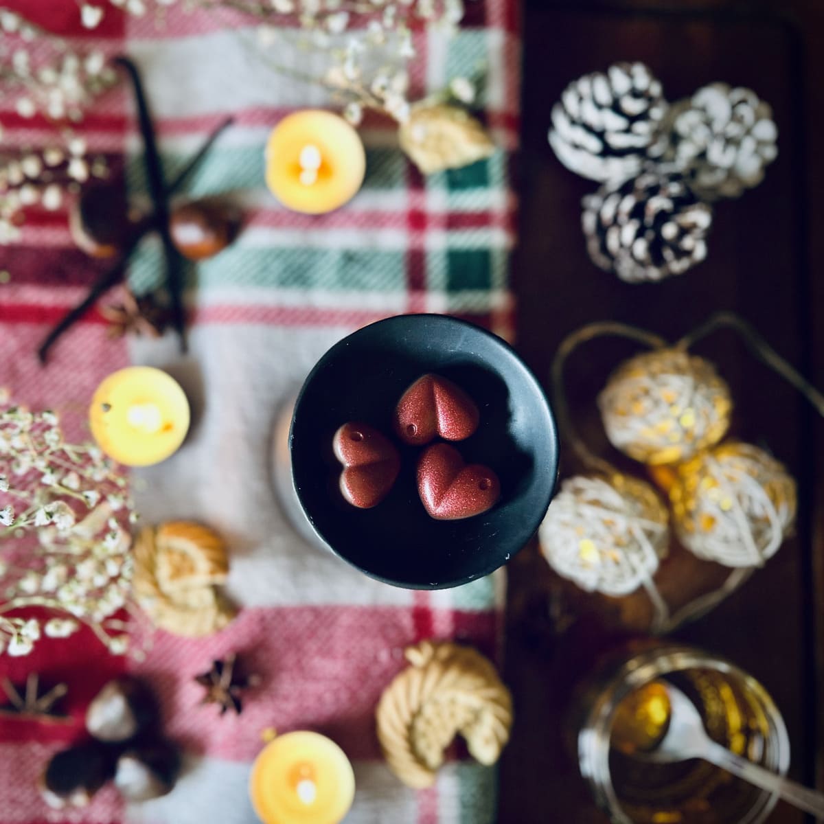 Three Red coloured Christmas Cookie scented heart shaped wax melts in a black wax melt burner on a festive tablecloth surrounded by chestnuts, vanilla, biscuits and caramel.